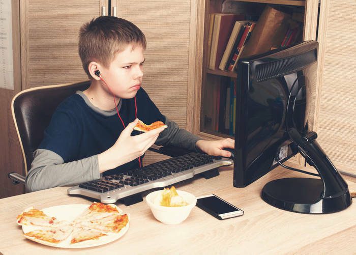 Kid eating pizza and surfing on internet or watshing video on PS. Boy in headphones eating fast food while using computer in his room.