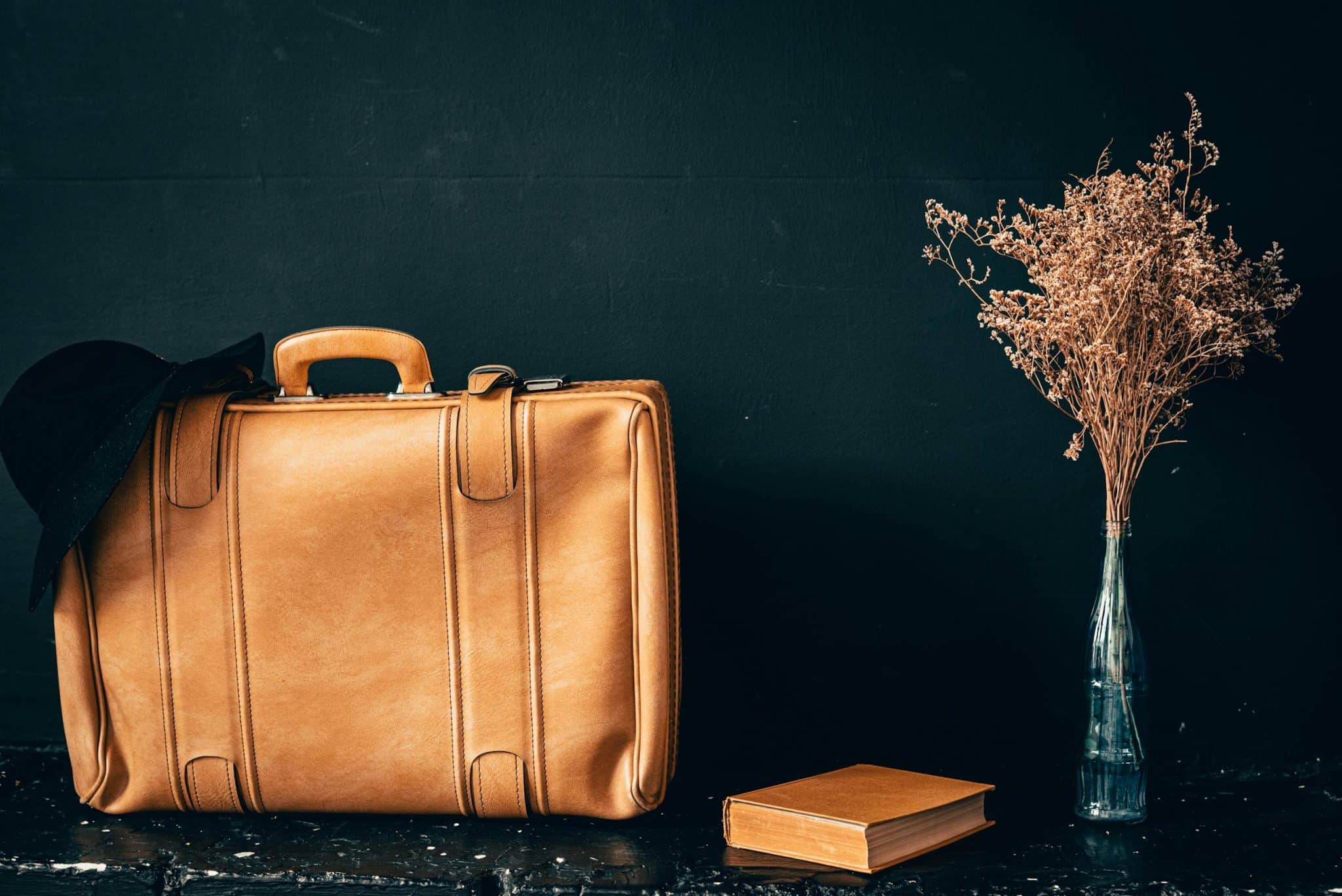A vintage leather suitcase paired with dried flowers in a vase and a book on a dark background.