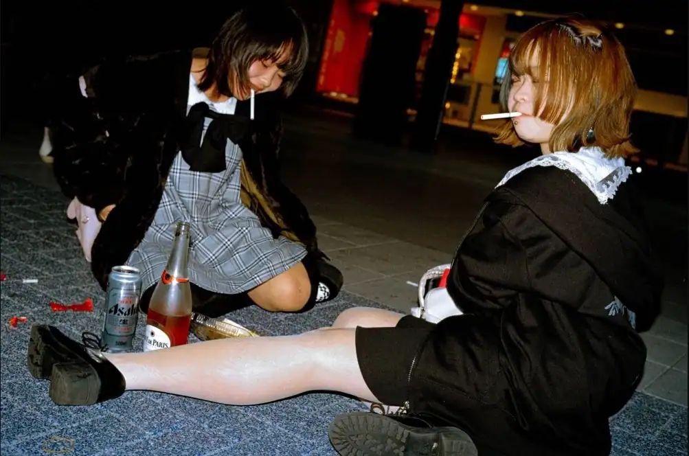 Two young girls sitting on the floor, one with a cigarette, in an urban setting.